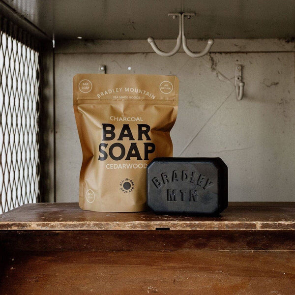 A bar of soap and its packaging, branded as 'BRADLEY MOUNTAIN CHARCOAL BAR SOAP', are placed on a wooden surface with a metal grid in the background. The packaging is brown with the brand name and product description printed on it.
