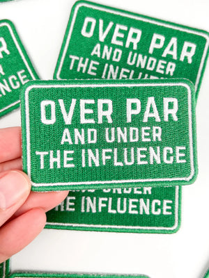Green patches with white text 'Over Par and Under the Influence' held by a hand.