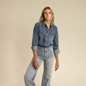 Woman wearing a blue denim shirt with pearl snap and jeans against a beige background