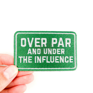 Green patch with text 'OVER PAR AND UNDER THE INFLUENCE' held by a hand on a white background