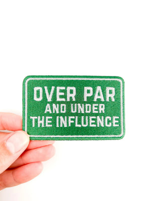 Green patch with text 'OVER PAR AND UNDER THE INFLUENCE' held by a hand on a white background