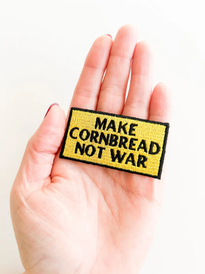 Hand holding a patch with 'MAKE CORNBREAD NOT WAR' text on a white background