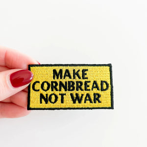 Patch with 'Make Cornbread Not War' text held by a hand on a white background
