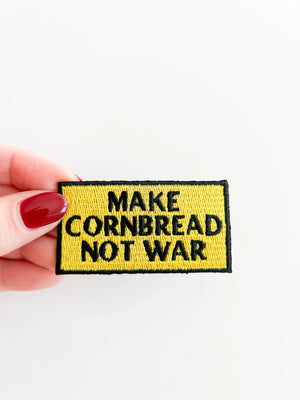 Patch with 'Make Cornbread Not War' text held by a hand on a white background