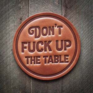 A brown leather coaster with the inscription 'DON'T FUCK UP THE TABLE' embossed on it, placed on a wooden surface.