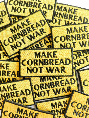 Yellow patches with 'MAKE CORNBREAD NOT WAR' in black block text on a pile.