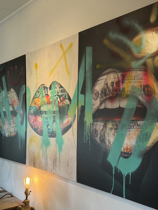 Image of a wall with alternating black and white canvases of open mouths with lips collaged with images of money, grafiti, and $100 notes. Nomad is spray painted across in blue lettering 