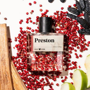 Perfume bottle labeled 'Preston' by Guy Fox surrounded by red berries and apples on a white background